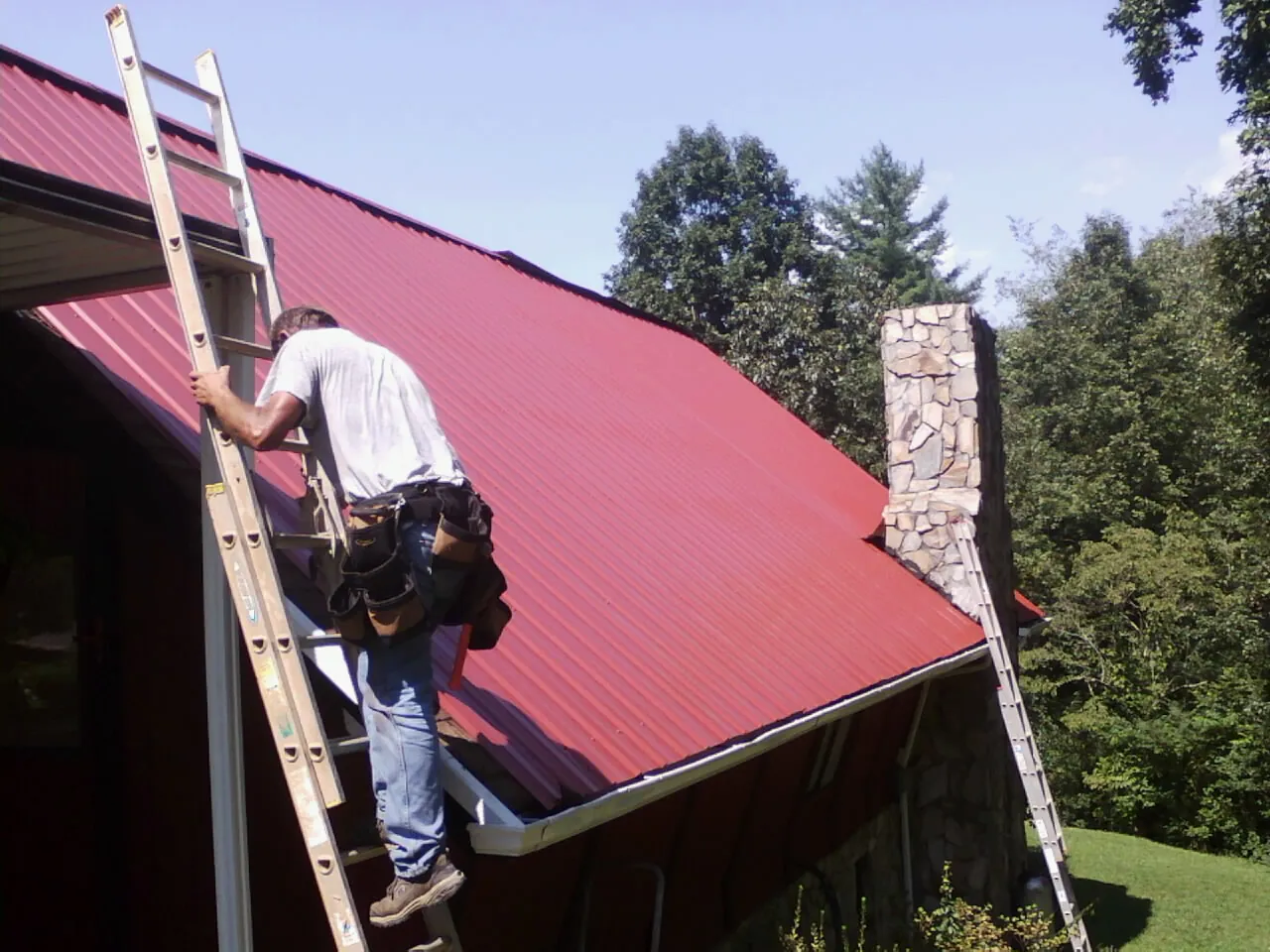 Roofer on a ladder beside a red metal roof and stone chimney — Soffit & Fascia Repair by Roofing Brothers in Eldora